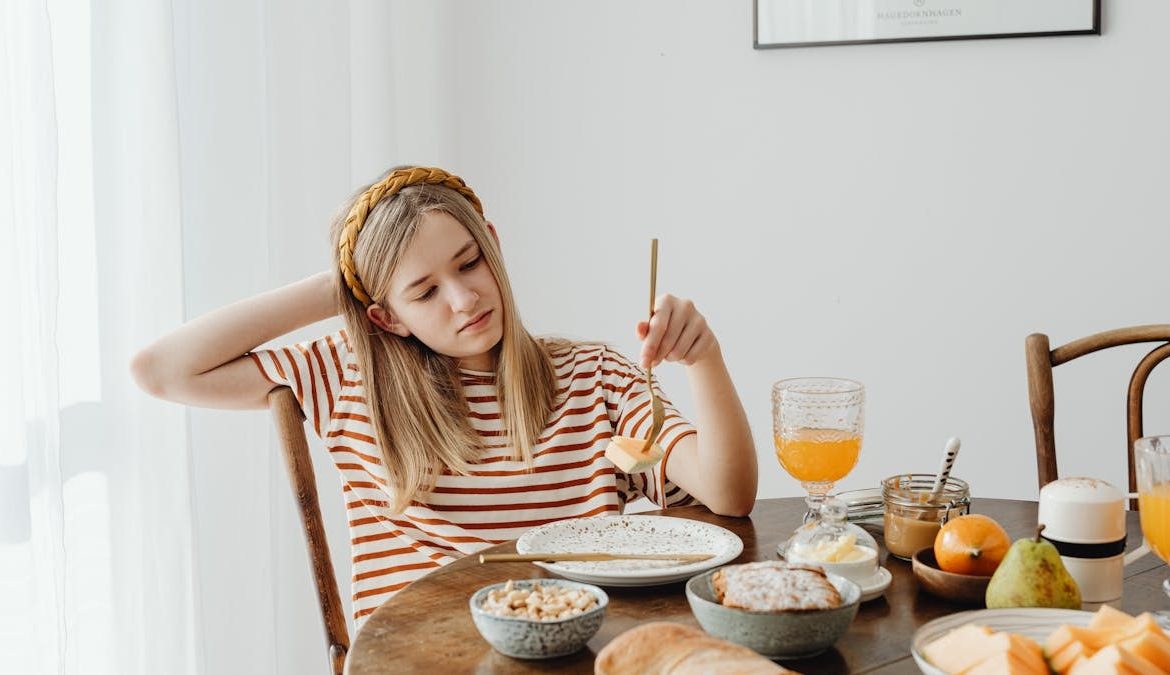 Teen girl in striped shirt at breakfast table with fruits, bread, and juice. Casual morning setting.